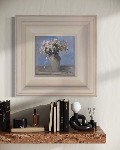 Small framed floral oil painting displayed above a wooden console table with books and sculptural decor in a serene contemporary interior.