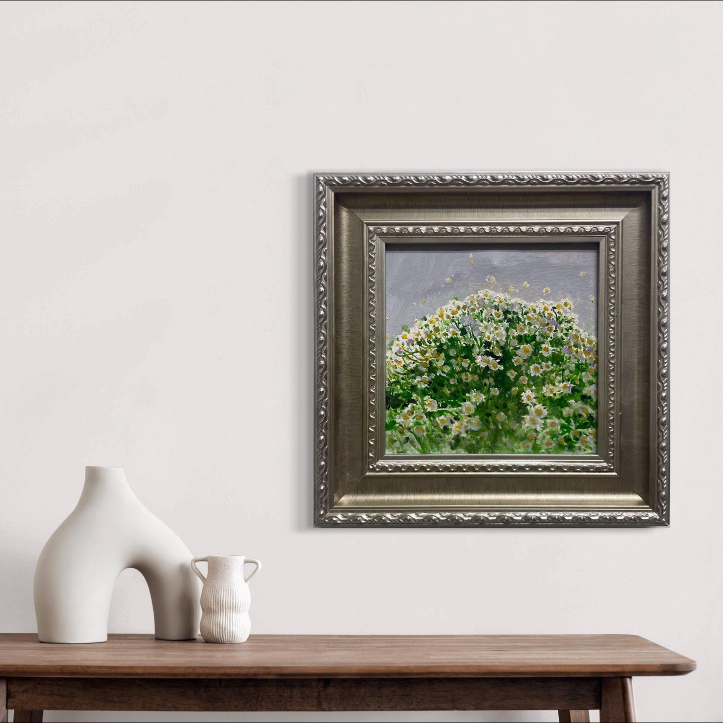 A small framed flower oil painting displayed above a wooden shelf with ceramic objects, bringing warmth, softness, and quiet botanical character into the modern interior.